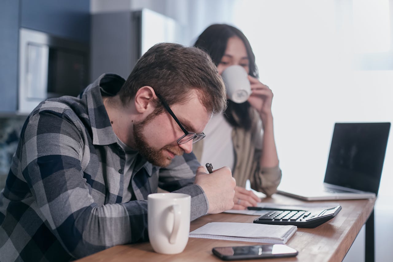 A couple sits at a kitchen table, reviewing finances with a laptop and calculator.